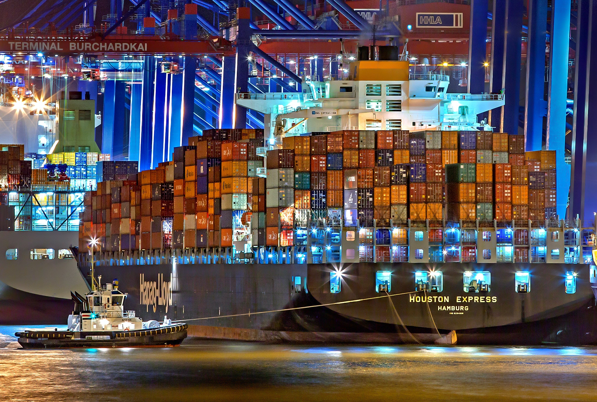 Ein großes, vollbeladenes Containerschiff der Hapag-Lloyd wird im Hamburger Hafen bei Nacht von einem Schlepper vom Kai weggezogen. Im Hintergrund befinden sich die blau-roten Hafenkräne der Containerterminals sowie ein weiteres Containerschiff.